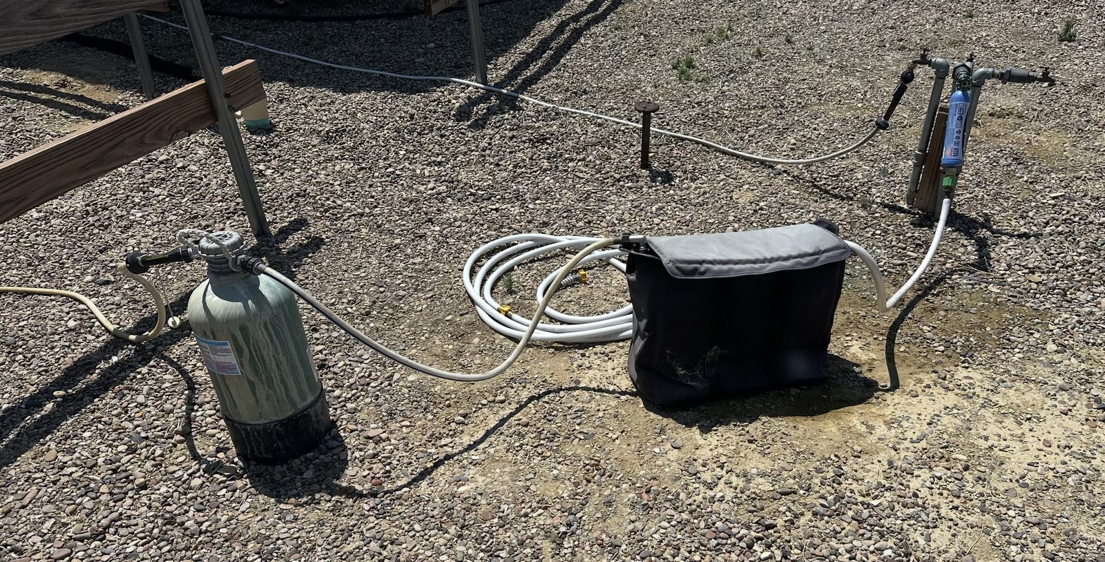 A portable RV water setup on the gravel at a campsite: a drag-out softener tank, a coil of hoses, a portable deionizer bag and an inline filter, all hooked up by hand at the spigot