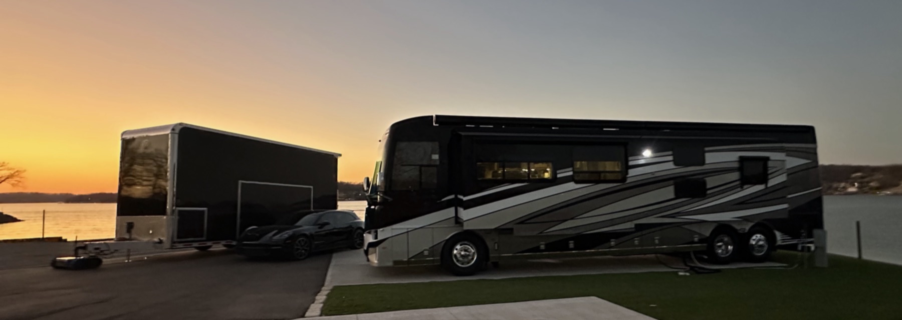 Luxury Newmar Dutch Star motorhome with an enclosed stacker trailer and sports car at a lakeside sunset